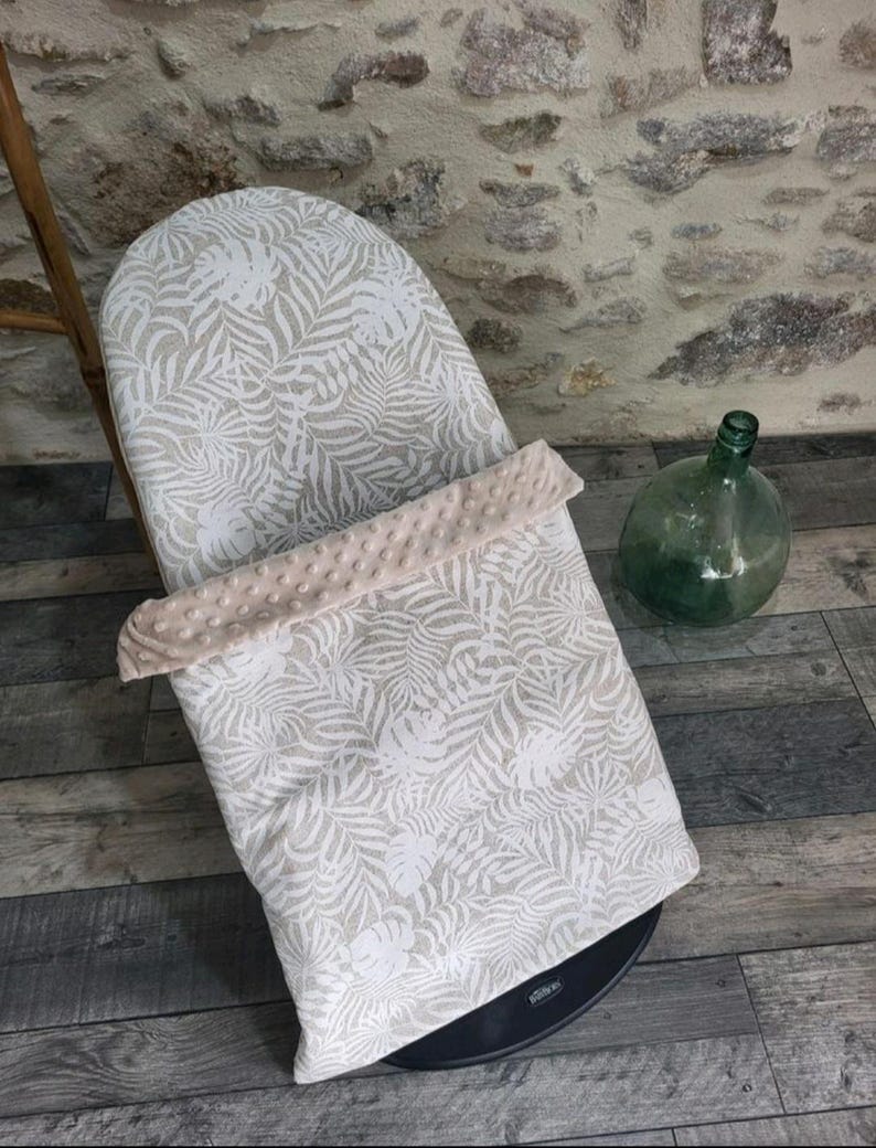 May include: A baby bouncer with a white and beige tropical leaf pattern. The bouncer has a soft, plush, beige border. A green glass bottle sits nearby on a wooden floor.