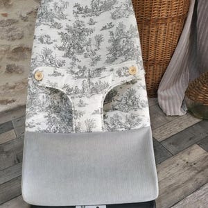 Pode incluir: Uma cadeira de balan&ccedil;o para beb&eacute; com um padr&atilde;o toile de Jouy a preto e branco na parte superior e uma sec&ccedil;&atilde;o inferior de veludo cinzento. A cadeira de balan&ccedil;o tem dois bot&otilde;es de madeira e uma base preta. O tecido apresenta uma cena pastoral.