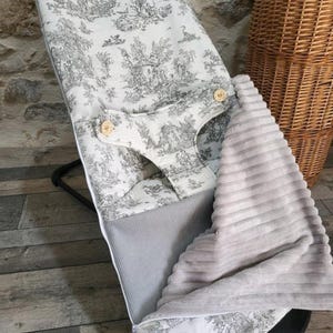 K&ouml;nnte beinhalten: Eine Babywippe mit schwarzem Rahmen und einem Bezug aus wei&szlig;em und grauem Toile-de-Jouy-Stoff. Der Bezug zeigt einen pastoralen Szenenaufdruck und Holzknopfverschl&uuml;sse. Ein grauer Rippstoff und eine weiche, graue Decke sind ebenfalls sichtbar.