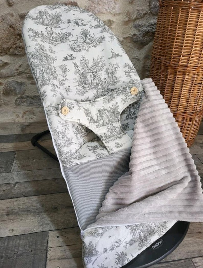 K&ouml;nnte beinhalten: Eine Babywippe mit schwarzem Rahmen und einem Bezug aus wei&szlig;em und grauem Toile-de-Jouy-Stoff. Der Bezug zeigt einen pastoralen Szenenaufdruck und Holzknopfverschl&uuml;sse. Ein grauer Rippstoff und eine weiche, graue Decke sind ebenfalls sichtbar.