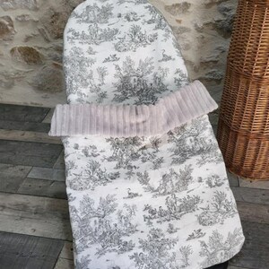 K&ouml;nnte beinhalten: Ein Baby-Sitzbezug mit einem schwarz-wei&szlig;en Toile-de-Jouy-Muster. Der Bezug hat eine weiche, hellrosa, plissierte Bord&uuml;re. Der Sitzbezug befindet sich auf einer schwarzen Basis, mit einem Holzboden und einem Weidenkorb im Hintergrund.