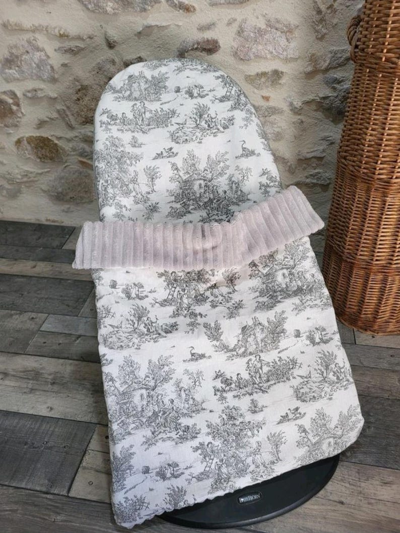 K&ouml;nnte beinhalten: Ein Baby-Sitzbezug mit einem schwarz-wei&szlig;en Toile-de-Jouy-Muster. Der Bezug hat eine weiche, hellrosa, plissierte Bord&uuml;re. Der Sitzbezug befindet sich auf einer schwarzen Basis, mit einem Holzboden und einem Weidenkorb im Hintergrund.