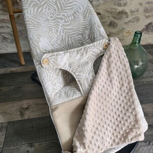 May include: A baby bouncer with a beige and white tropical leaf pattern. The bouncer has a soft, textured blanket and wooden button details. The bouncer is designed for infant comfort and support.