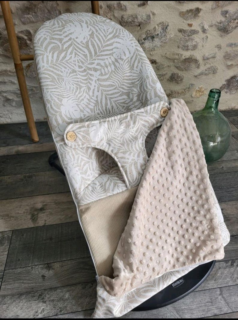 May include: A baby bouncer with a beige and white tropical leaf pattern. The bouncer has a soft, textured blanket and wooden button details. The bouncer is designed for infant comfort and support.