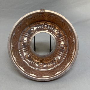 May include: A copper-colored bundt cake pan with a decorative, circular design. The pan has a central hole and a series of embossed patterns, including circles and curved lines. A small loop is attached at the top for hanging.