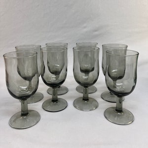 May include: A set of eight gray-tinted wine glasses. Each glass has a rounded bowl, a slender stem, and a circular base. The glasses are arranged in a group, showcasing their uniform design and color. The glassware is suitable for serving wine.