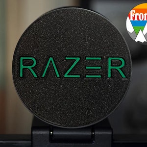 Peut inclure: Un capuchon d'objectif d'appareil photo rond noir avec le mot "RAZER" en lettres vertes.