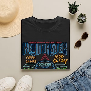 May include: A black t-shirt featuring a neon-style graphic with the text "THERE'S NO GATE WE CAN'T OPEN", "KEYMASTER", "OPEN 24 HRS", "555-2368", and "Lock in Key" in blue, red, yellow, and green colours against the dark fabric background.