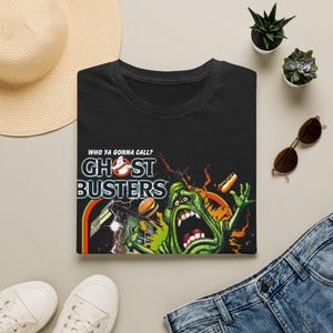Peut inclure: Un t-shirt noir avec une impression graphique du logo Ghostbusters et le texte "WHO YA GONNA CALL?" au-dessus. Le motif comprend une créature verte à la bouche grande ouverte, un hamburger, un hot-dog et un bâtiment avec des éclairs.