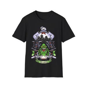 Peut inclure: Un t-shirt noir à manches courtes avec une impression graphique représentant un personnage en guimauve blanc, un fantôme vert, deux créatures noires à cornes et une silhouette urbaine. Sous l'illustration, une bannière verte affiche WHO YOU GONNA CALL.