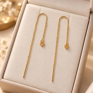 May include: A pair of gold-toned threader earrings featuring a delicate chain design. Each earring consists of a thin metal wire that loops through the ear, ending in a straight post on one side and a small, dangling heart-shaped charm on the other.