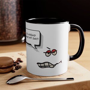 Puede incluir: Taza de cerámica blanca con interior y asa negros. La taza presenta una cara de dibujos animados con el texto "GO AHEAD MAKE MY DAY!" en un bocadillo. La cara tiene ojos con bordes rojos y una boca que hace muecas.
