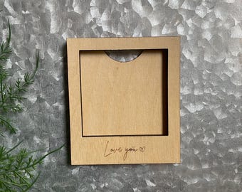 May include: A square, wooden card holder with a rectangular opening. The holder is light brown with the words "Love you" and a heart etched into the wood. The background is a textured, metallic surface.