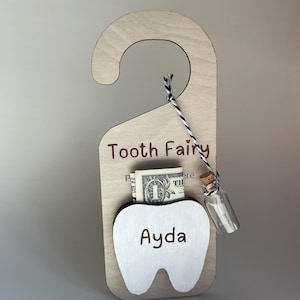 May include: Wooden tooth fairy door hanger with a white tooth cutout, the name "Ayda" is engraved on the tooth. A dollar bill and a small glass bottle are tucked into the tooth cutout. The door hanger has the text "Tooth Fairy" engraved on it.