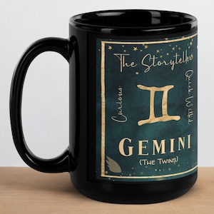 May include: Black ceramic mug with a gold design featuring the Gemini zodiac sign, the text "The Storyteller", "Curious", "Quick-Witted", and "GEMINI (THE TWINS)".