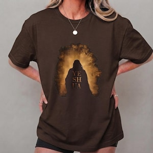 Puede incluir: Una camiseta de manga corta de color marrón oscuro con un gráfico central de una figura silueteada sobre un fondo brillante de nubes de color dorado. El texto "YESHUA" está impreso en formato vertical y apilado sobre el pecho.