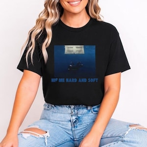 Puede incluir: Camiseta negra con un gráfico azul de una persona bajo el agua mirando hacia un edificio. El texto "HI ME HARD AND SOFT" está impreso en azul debajo. La persona lleva vaqueros azules.
