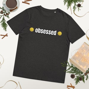 May include: A dark heather grey t-shirt with the text "obsessed" printed in white. Two yellow pickleball paddles are on either side of the text.
