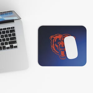 May include: A rectangular mouse pad with a dark blue gradient background featuring an orange bear head logo in the center. A white computer mouse rests on the right side of the pad, positioned next to a silver laptop with a black keyboard.