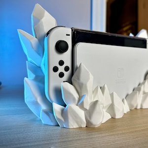 May include: A white Nintendo Switch console is displayed in a white crystal-shaped stand. The console is in its docked position with the Joy-Con controllers attached. The stand is made of 3D printed plastic and resembles a cluster of crystals.