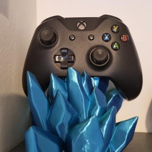 May include: A black Xbox controller sits on a blue, 3D printed stand shaped like ice crystals.