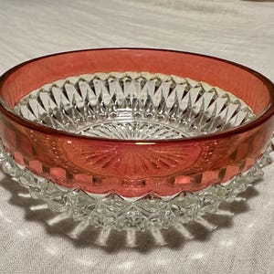 May include: A clear glass bowl with a ruby red rim and accents. The bowl features a diamond-cut pattern on the interior and a textured base. The bowl is likely vintage and suitable for serving or display.