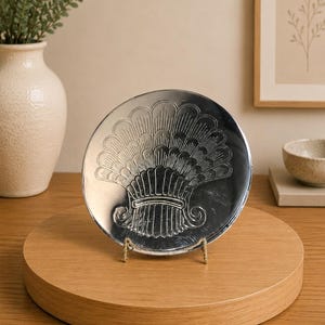 May include: A silver-colored decorative plate with an embossed fan design, displayed on a wooden stand. The plate is round and features intricate detailing. The stand is made of light-colored wood, and the background includes a vase and framed artwork.