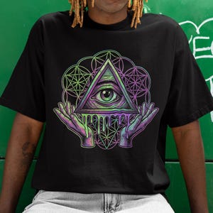 Puede incluir: Camiseta negra con un gráfico colorido de un ojo dentro de un triángulo, sostenido por manos, con formas geométricas. El diseño tiene tonos verdes, morados y rosas. La persona lleva vaqueros de color claro.