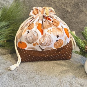 May include: A drawstring bag featuring a fall-themed pumpkin pattern. The bag has a white background with orange, brown, and tan pumpkins. The bottom of the bag is brown with white star-shaped dots. The drawstring is a thick, off-white rope.