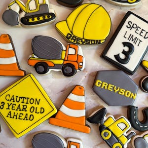 May include: Assortment of construction-themed cookies, including a yellow hard hat, cement trucks, and road signs. The cookies are decorated with black, orange, and gray icing. One sign reads "CAUTION 3 YEAR OLD AHEAD."