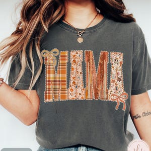 May include: Dark gray t-shirt with the word "MAMA" in a decorative, fall-themed design. The letters feature plaid, floral, and wood-grain patterns, with decorative bows. The shirt is a relaxed fit.