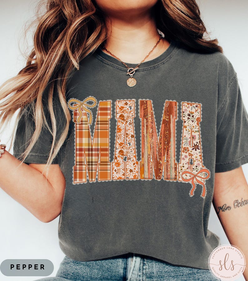 May include: Dark gray t-shirt with the word "MAMA" in a decorative, fall-themed design. The letters feature plaid, floral, and wood-grain patterns, with decorative bows. The shirt is a relaxed fit.