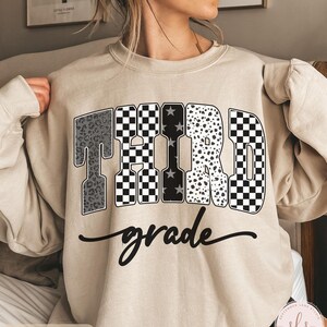 May include: Sand-colored sweatshirt with the words "THIRD grade" in a bold, decorative font. The letters feature various patterns, including leopard print, checkered, and star designs. The word "grade" is written in a cursive script below the word "THIRD".