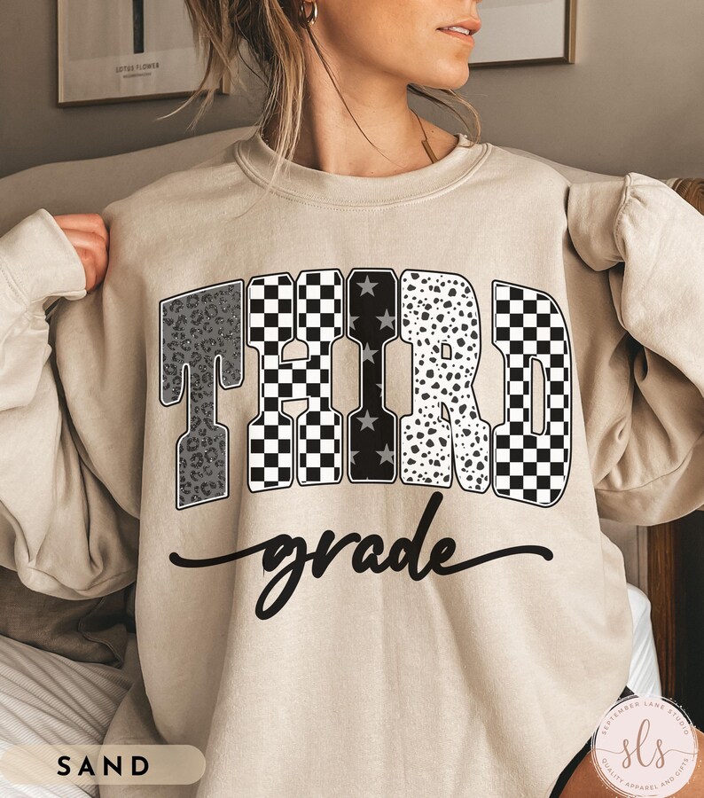 May include: Sand-colored sweatshirt with the words "THIRD grade" in a bold, decorative font. The letters feature various patterns, including leopard print, checkered, and star designs. The word "grade" is written in a cursive script below the word "THIRD".
