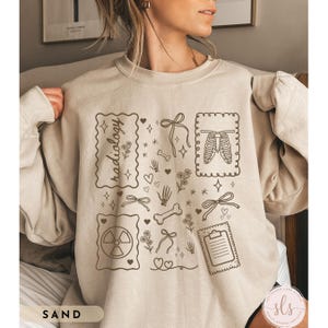 Puede incluir: Sudadera de color arena con un dise&ntilde;o gr&aacute;fico beige que presenta la palabra "radiology" en un marco decorativo, junto con ilustraciones de huesos, lazos, corazones y una caja tor&aacute;cica. La palabra "SAND" est&aacute; impresa en la parte inferior izquierda.
