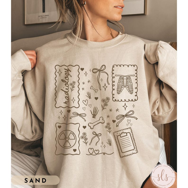 Puede incluir: Sudadera de color arena con un dise&ntilde;o gr&aacute;fico beige que presenta la palabra "radiology" en un marco decorativo, junto con ilustraciones de huesos, lazos, corazones y una caja tor&aacute;cica. La palabra "SAND" est&aacute; impresa en la parte inferior izquierda.