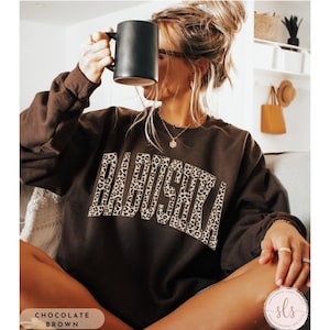May include: Chocolate brown sweatshirt with the word "BABUSHKA" in a leopard print design. The model is holding a black mug. The sweatshirt is part of the "September Lane Studio" collection.