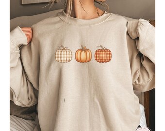 May include: A sand-coloured sweatshirt featuring three watercolour pumpkins in shades of orange and brown, one with a plaid pattern. The word "SAND" is printed at the bottom. The sweatshirt has long sleeves.