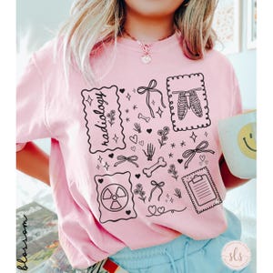 Puede incluir: Una camiseta rosa claro con un dise&ntilde;o gr&aacute;fico negro que incluye la palabra "radiolog&iacute;a", huesos, lazos, flores y un s&iacute;mbolo de radiaci&oacute;n. La camiseta tiene cuello redondo y mangas cortas.