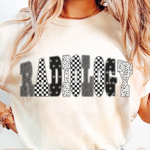 K&ouml;nnte beinhalten: Elfenbeinfarbenes T-Shirt mit dem Wort "RADIOLOGY" in einem auff&auml;lligen, gemusterten Design. Die Buchstaben weisen eine Mischung aus schwarz-wei&szlig; karierten, Stern- und Tiermuster auf. Das Shirt hat einen Rundhalsausschnitt.