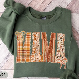 May include: Military green sweatshirt with the word "MAMA" in a decorative, fall-themed design. The letters feature plaid, floral, and textured patterns in orange, brown, and cream colors. The sweatshirt has a crew neck and long sleeves.