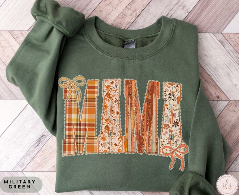 May include: Military green sweatshirt with the word "MAMA" in a decorative, fall-themed design. The letters feature plaid, floral, and textured patterns in orange, brown, and cream colors. The sweatshirt has a crew neck and long sleeves.