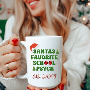 May include: White ceramic mug with a green and red design that says "Santa's Favorite School Psych" with a red ornament and a green Christmas tree. The mug also has the name "Ms. Janet" written in red.