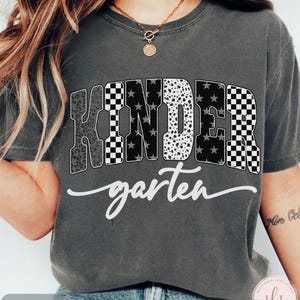May include: A dark gray t-shirt with the word "KINDERgarten" in a mix of patterns, including leopard print, checkered, and stars. The word "garten" is in a cursive font. The shirt has a crew neck and short sleeves.