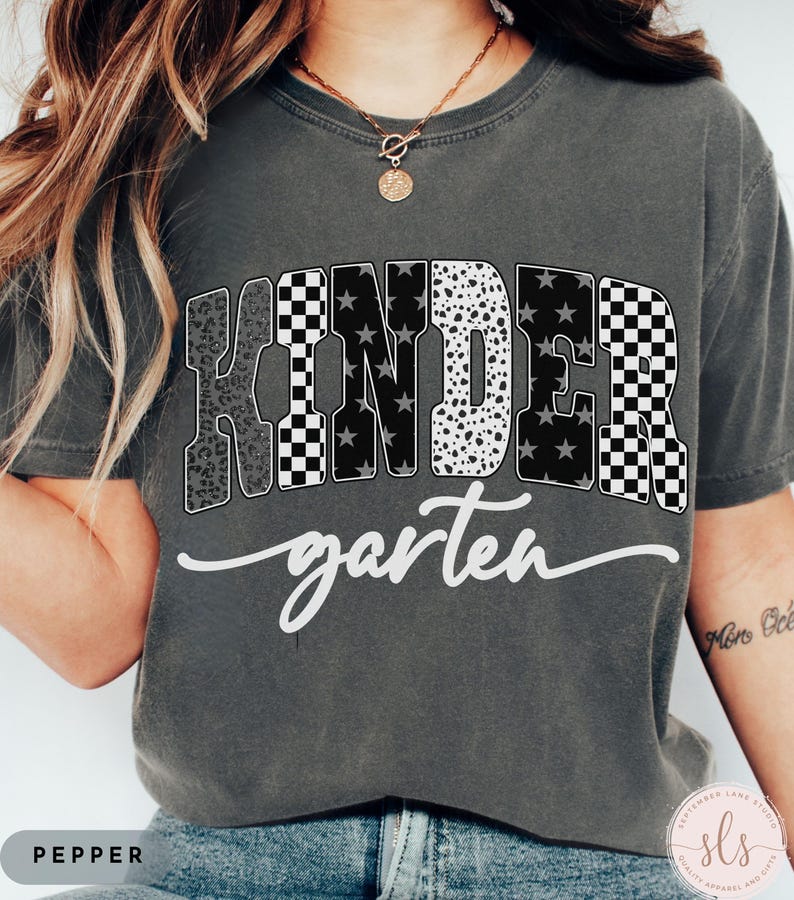 May include: A dark gray t-shirt with the word "KINDERgarten" in a mix of patterns, including leopard print, checkered, and stars. The word "garten" is in a cursive font. The shirt has a crew neck and short sleeves.