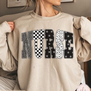 May include: Sand-colored sweatshirt with the word "NURSE" in a bold, multi-patterned design. The letters feature leopard print, checkered, star, and polka dot patterns. The sweatshirt is a neutral color and has a crew neck.