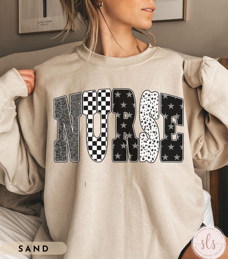 May include: Sand-colored sweatshirt with the word "NURSE" in a bold, multi-patterned design. The letters feature leopard print, checkered, star, and polka dot patterns. The sweatshirt is a neutral color and has a crew neck.