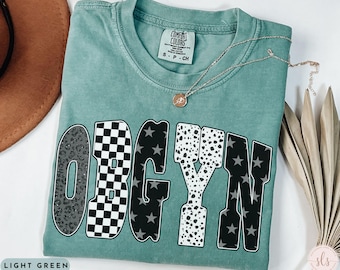 May include: Light green t-shirt with the word "OBGYN" in a bold, multi-patterned design. The letters feature leopard print, a checkerboard pattern, black stars, and dalmatian spots. A gold necklace with a pendant rests on the shirt.