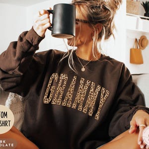 Könnte beinhalten: Dunkelbraunes, individuelles Sweatshirt mit dem Wort "GRANNY" im Leopardenmuster. Das Sweatshirt hat lange Ärmel und einen Rundhalsausschnitt. Die Wörter "CUSTOM SWEATSHIRT" und "DARK CHOCOLATE" sind ebenfalls sichtbar.