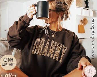 Puede incluir: Sudadera personalizada de color chocolate oscuro con la palabra "GRANNY" en estampado de leopardo. La sudadera tiene mangas largas y cuello redondo. También se ven las palabras "CUSTOM SWEATSHIRT" y "DARK CHOCOLATE".